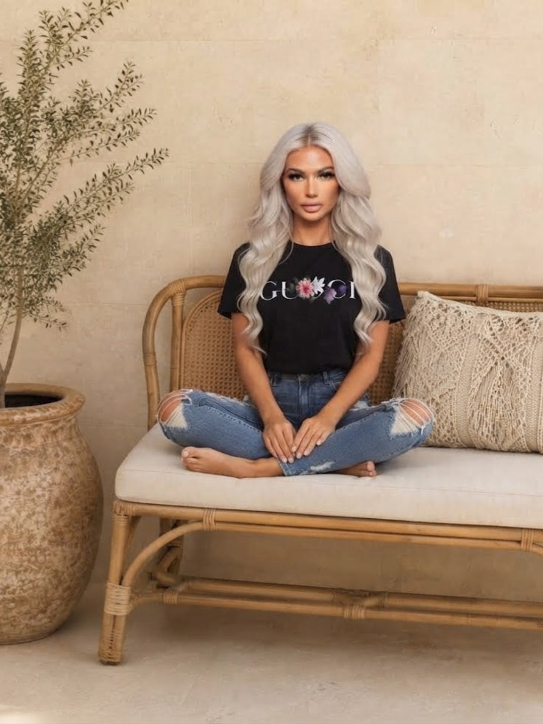 A woman with long silver hair sits cross-legged on a rattan bench wearing a Villa Vibes black t-shirt with a colorful 'GUCCI' logo and distressed blue jeans, set against a warm beige wall with a potted plant.