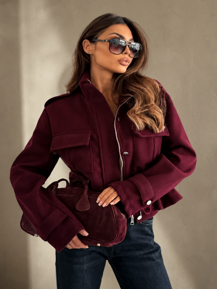 A woman wearing a Villa Vibes Blaise cropped burgundy jacket with silver zipper details, dark sunglasses, and dark blue jeans, holding a matching burgundy handbag against a textured beige wall.