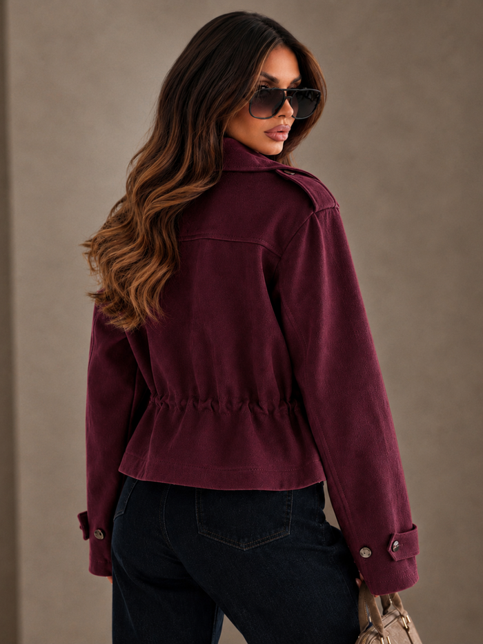 A woman wearing a Villa Vibes Blaise cropped, long-sleeve burgundy jacket with a gathered waist and dark jeans, photographed from a side-back angle against a neutral textured beige background