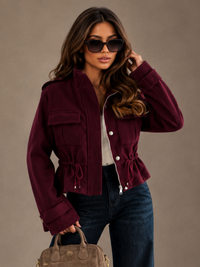 A woman models a Villa Vibes Blaise cropped burgundy jacket with a drawstring waist, paired with dark wide-leg jeans, a white top, and a taupe handbag against a neutral studio backdrop.