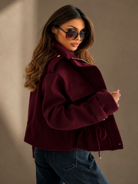 A woman modeling a Villa Vibes Blaise cropped, oversized deep burgundy jacket with a drawstring waist, paired with dark blue jeans and sunglasses, set against a warm beige studio background.