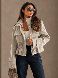 A woman wearing a Villa Vibes Blaise light beige cropped utility jacket with a high collar, button front, and drawstring hem, styled with dark blue jeans, a white top, and sunglasses against a neutral beige background.