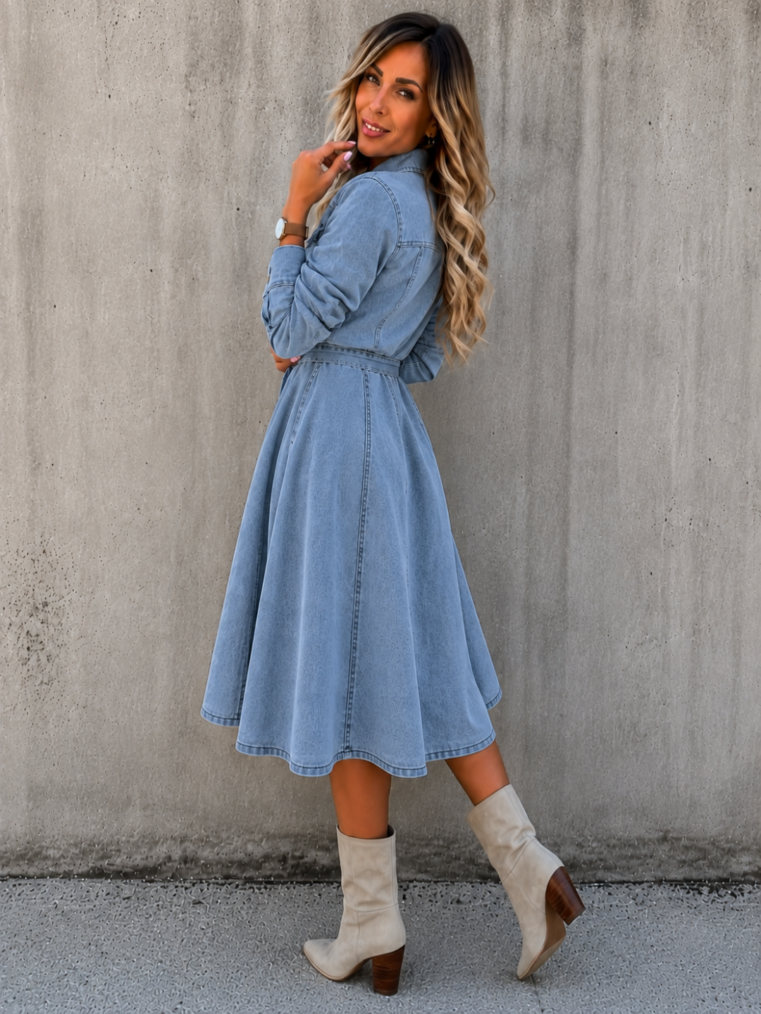 A woman wearing a Villa Vibes Blue Belle Chambray light blue long-sleeve midi dress with a tie waist, paired with beige ankle boots, posing against a textured concrete wall.