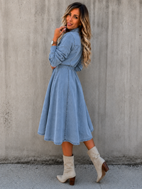A woman wearing a Villa Vibes Blue Belle Chambray light blue long-sleeve midi dress with a tie waist, paired with beige ankle boots, posing against a textured concrete wall.