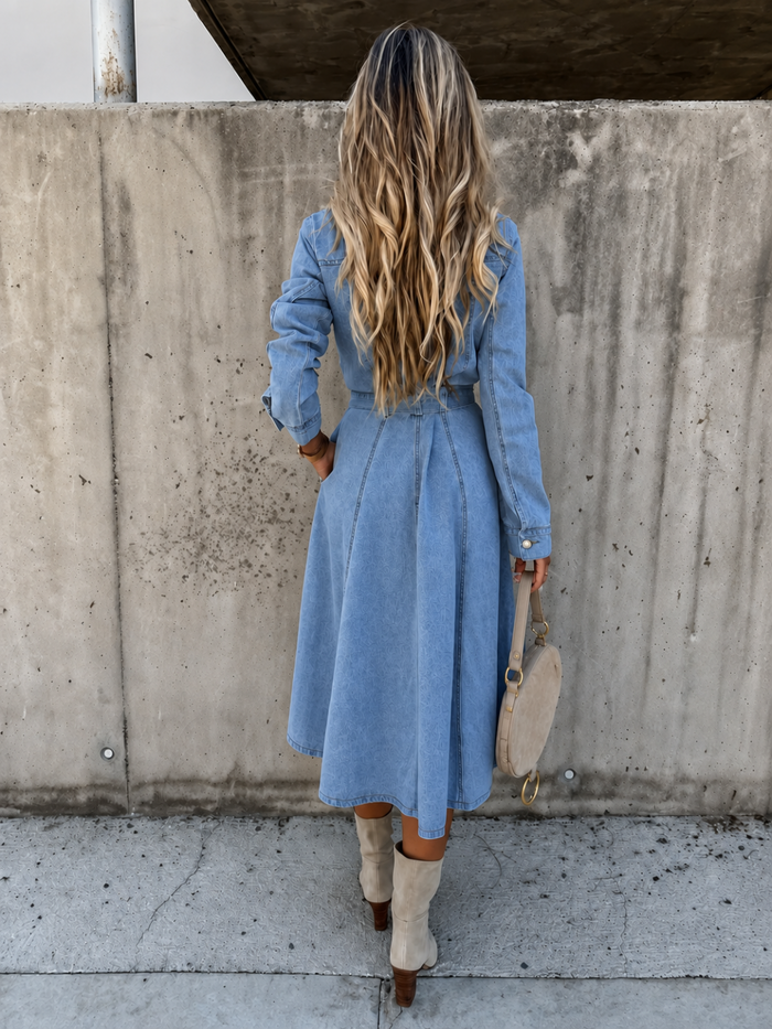 A woman viewed from behind wearing a Villa Vibes Blue Belle light blue chambray shirt dress with long sleeves and a tie waist, paired with beige knee-high boots and holding a beige handbag, standing against a concrete wall.