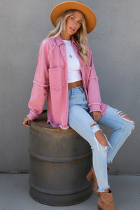 Woman wearing a Villa Vibes pink jacket and ripped jeans sitting on a barrel against a gray wall.