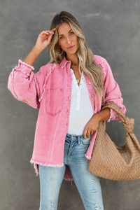 Woman wearing a Villa Vibes Brydie pink denim jacket, white shirt, and blue jeans, holding a woven bag against a gray background.