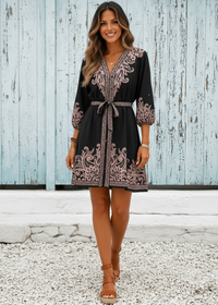 Woman wearing a Villa Vibes Cassandra black dress with intricate patterns standing against a wooden wall.