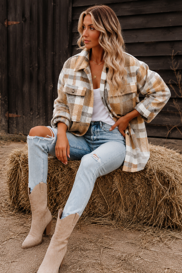 Woman sitting on a hay bale wearing a Villa Vibes Cassidy plaid shirt, jeans, and boots in front of a wooden wall.