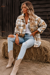 Woman sitting on a hay bale wearing a Villa Vibes Cassidy plaid shirt, jeans, and boots in front of a wooden wall.