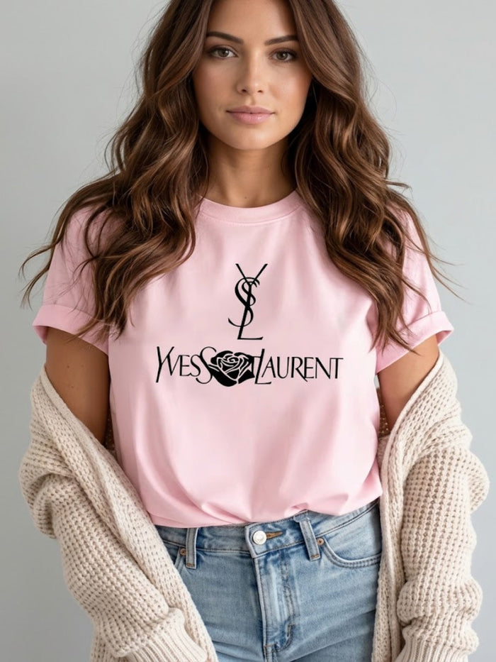 A woman wearing a Villa VIbes Designer Inspired light pink short-sleeve T-shirt with a black Yves Saint Laurent logo and rose graphic on the chest, styled with an off-white knit cardigan and blue jeans against a light gray background.