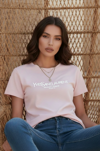 Woman wearing a Villa Vibes pink t-shirt with branded logo and blue jeans sitting against a woven wall.