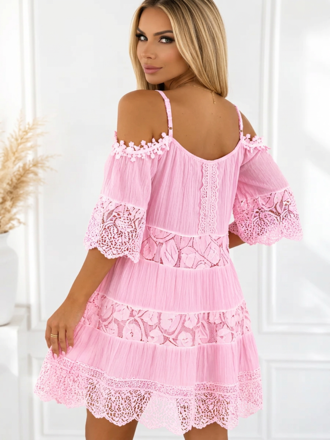 A woman wearing a Villa Vibes Fallon light pink cold-shoulder mini dress with lace trim and tiered ruffles, shown from the back in a bright indoor setting.