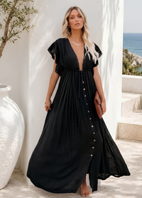 A woman models a Villa Vibes Holly black maxi dress with short flutter sleeves, a deep V-neckline, and a button-down front, set against a Mediterranean-style outdoor background with a white wall, large planter, and blue sea in the distance.