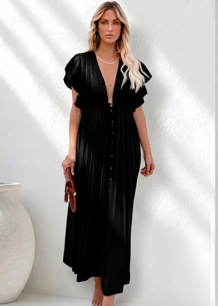 A woman stands barefoot in a bright white architectural setting with strong sunlight and shadows, wearing a Villa Vibes Holly black raw edge maxi dress with short puff sleeves, a deep V-neckline, and a button front. She holds a small brown clutch.