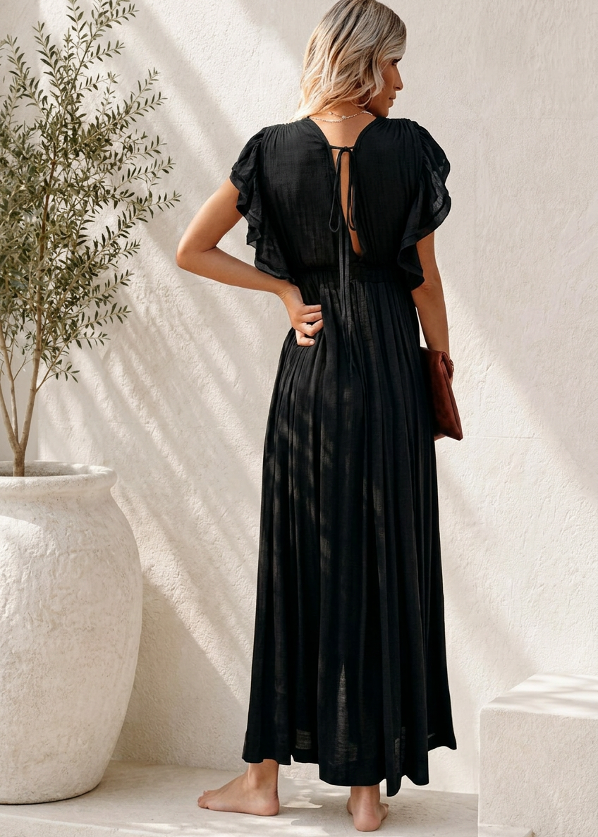 A woman stands barefoot in a minimalist indoor setting, wearing a Villa Vibes Holly black maxi dress with a sheer ruffled short sleeve and a tie-back neckline. She is posed with her back to the camera, holding a small brown handbag, next to a large white planter with a leafy plant.