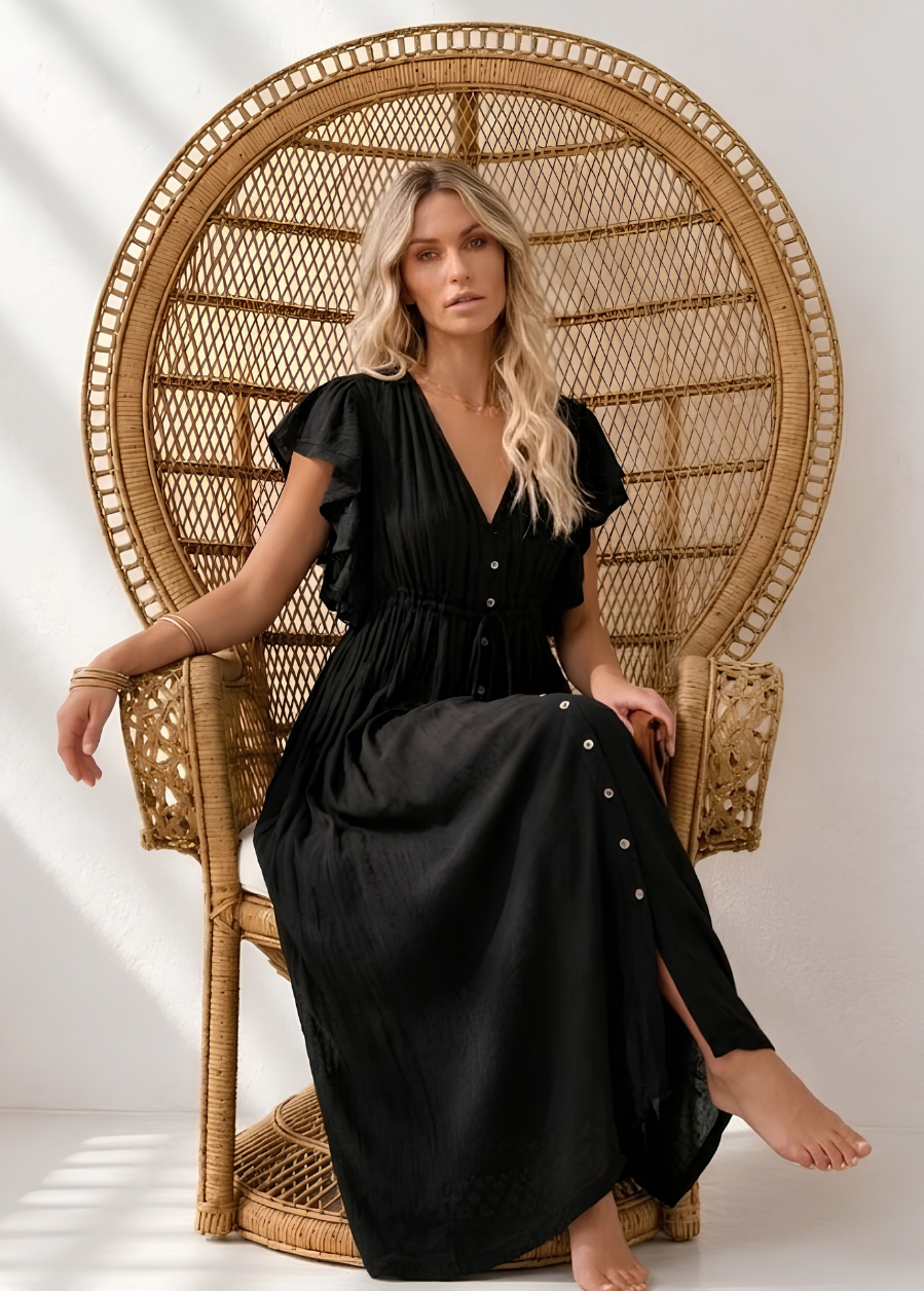 A woman wearing a Villa Vibes Holly black midi dress sits barefoot in a tall woven rattan peacock chair against a white wall with soft window shadows.
