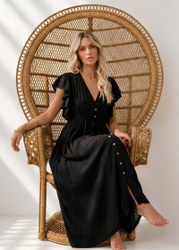 A woman wearing a Villa Vibes Holly black midi dress sits barefoot in a tall woven rattan peacock chair against a white wall with soft window shadows.