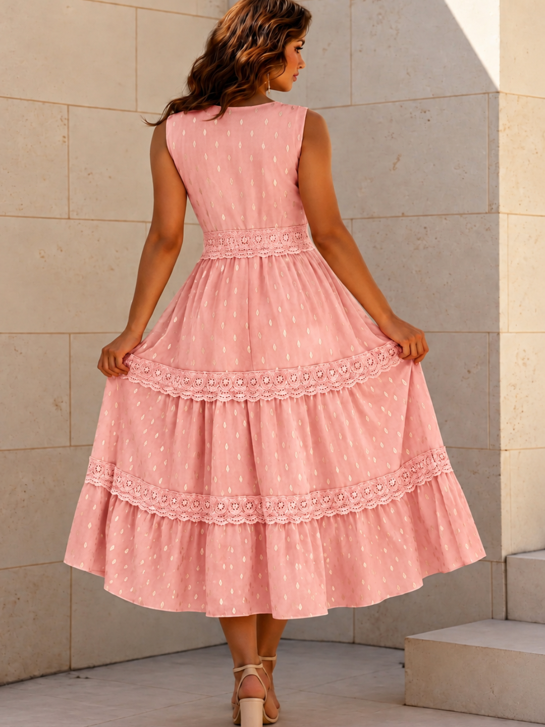 A woman wearing a Villa Vibes Indra sleeveless pink tiered midi dress with white polka dots and lace trim, shown from the back against a beige stone wall.