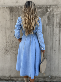 A woman viewed from behind wearing a Villa Vibes Isla light blue denim midi dress with long sleeves and a tie waist, standing against a textured concrete wall and holding a beige handbag.