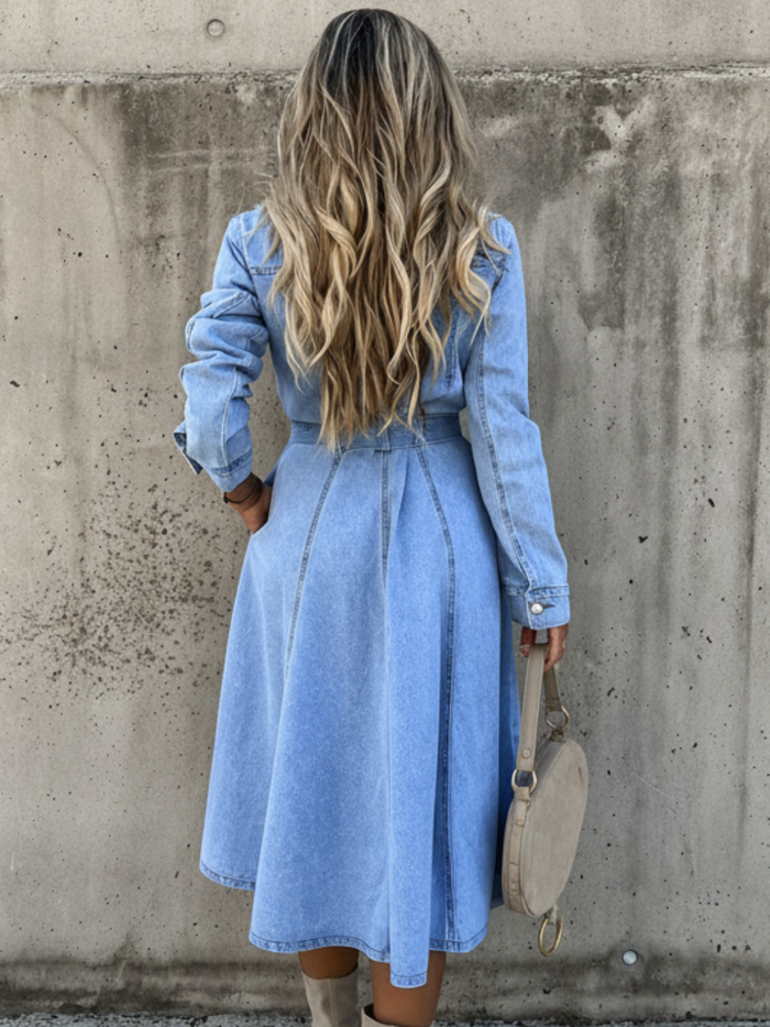 A woman viewed from behind wearing a Villa Vibes Isla light blue denim midi dress with long sleeves and a tie waist, standing against a textured concrete wall and holding a beige handbag.