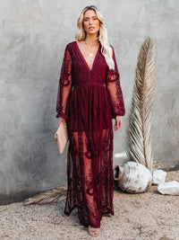 Woman wearing a Villa Vibes Janis burgundy lace dress against a gray wall with decorative elements.