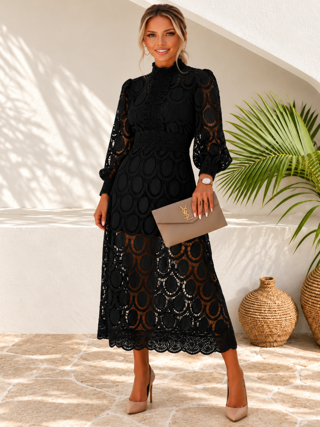 A woman models a Villa Vibes Jasmine black midi-length lace dress with a mock neck, long sleeves, and a matching waist belt. She is holding a taupe clutch and wearing beige heels, standing in a sunlit outdoor patio setting with a potted palm and woven vases.