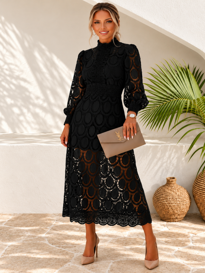 A woman models a Villa Vibes Jasmine black midi-length lace dress with a mock neck, long sleeves, and a matching waist belt. She is holding a taupe clutch and wearing beige heels, standing in a sunlit outdoor patio setting with a potted palm and woven vases.