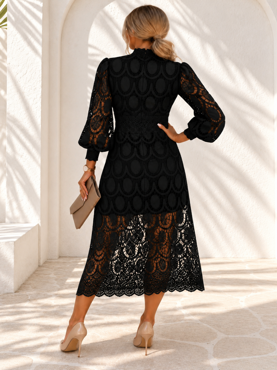 A woman viewed from behind wearing a Villa Vibes Jasmine black midi dress with a textured scalloped bodice and sheer lace sleeves, standing in a bright white arched corridor and holding a beige clutch.