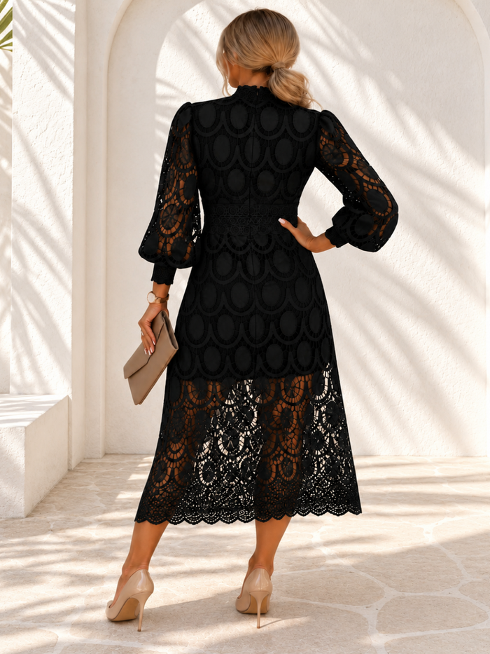 A woman viewed from behind wearing a Villa Vibes Jasmine black midi dress with a textured scalloped bodice and sheer lace sleeves, standing in a bright white arched corridor and holding a beige clutch.