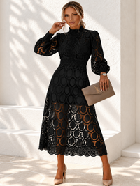 A woman models a Villa Vibes Jasmine black midi dress with long sheer lace sleeves, a high mock neck, and a textured circular lace overlay. She is holding a taupe clutch and wearing beige heels, standing against a white wall with soft shadows.