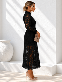 A woman in a Villa Vibes Jasmine black lace midi dress with a high neck and long sleeves stands in profile against a white textured wall, holding a beige clutch and wearing beige heels.