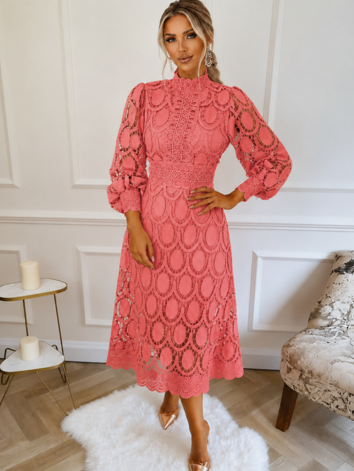 A woman models a Villa Vibes Jasmine coral pink lace midi dress with long balloon sleeves and a high mock neck in a bright room with white walls, a white rug, and a patterned armchair.