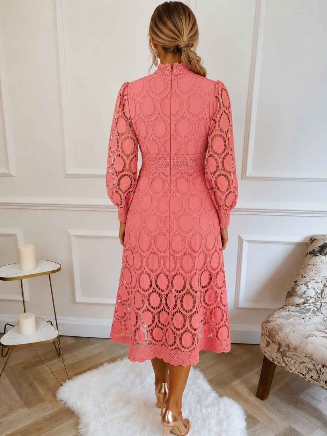 A woman viewed from behind wearing a VIlla Vibes coral pink lace midi dress with long sleeves and an allover circular lace pattern, standing in a bright room with white paneled walls, a white rug, and light wood flooring.
