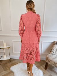 A woman viewed from behind wearing a VIlla Vibes coral pink lace midi dress with long sleeves and an allover circular lace pattern, standing in a bright room with white paneled walls, a white rug, and light wood flooring.
