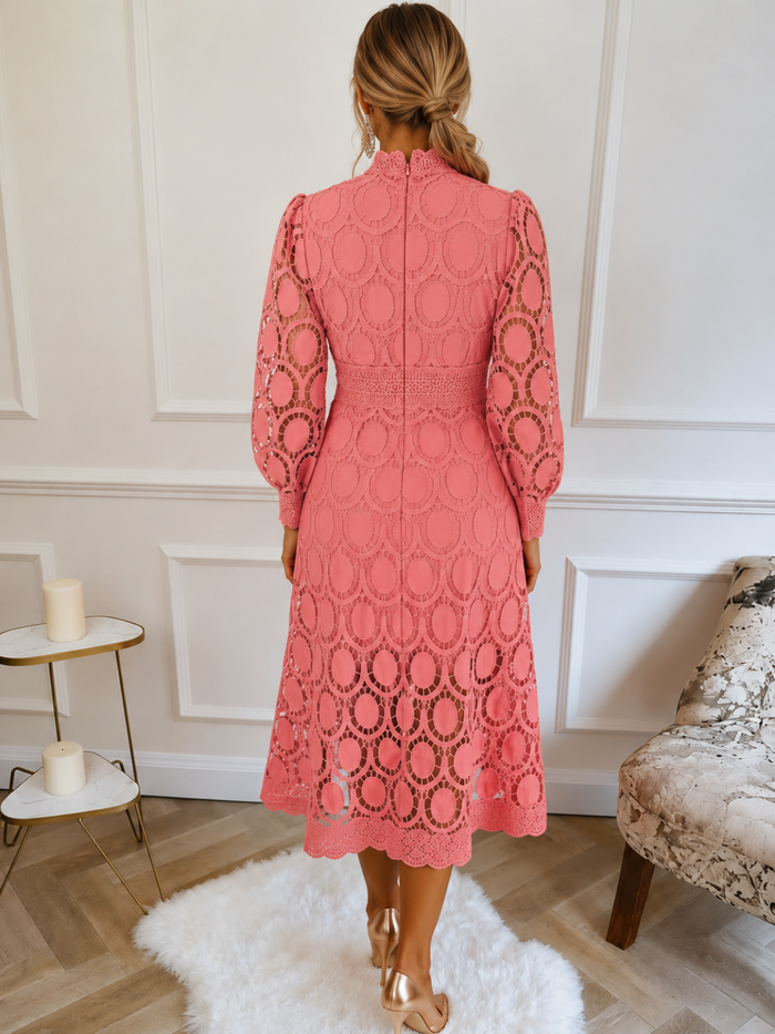 A woman viewed from behind wearing a VIlla Vibes coral pink lace midi dress with long sleeves and an allover circular lace pattern, standing in a bright room with white paneled walls, a white rug, and light wood flooring.