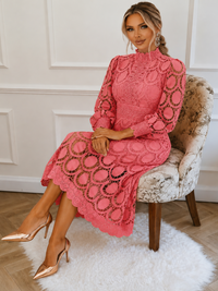 A woman sits on a patterned armchair wearing a Villa Vibes Jasmine pink lace midi dress with long sleeves and circular lace cutouts, paired with metallic rose gold heels in an indoor room with white paneled walls and a white rug.