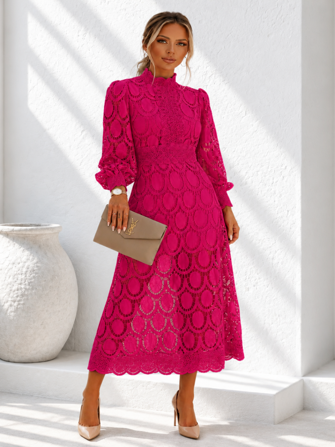 A woman models a Villa Vibes Jasmine bright magenta lace midi dress with long sleeves and a high mock neck, holding a taupe clutch bag. She is standing in a bright white room with architectural shadows.