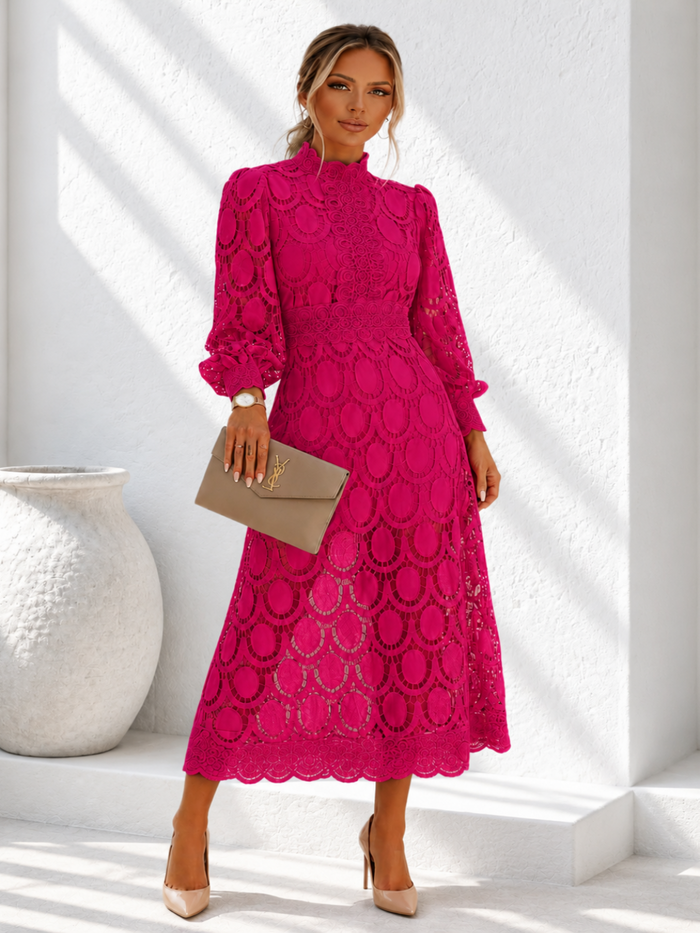 A woman models a Villa Vibes Jasmine bright magenta lace midi dress with long sleeves and a high mock neck, holding a taupe clutch bag. She is standing in a bright white room with architectural shadows.