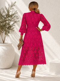 A woman shown from the back wearing a  Villa Vibes Jasmine bright fuchsia pink lace midi dress with long balloon sleeves and a high neckline. She is holding a small taupe clutch and wearing nude heels, standing in a sunlit room with a white textured wall, a potted plant, and a white block.