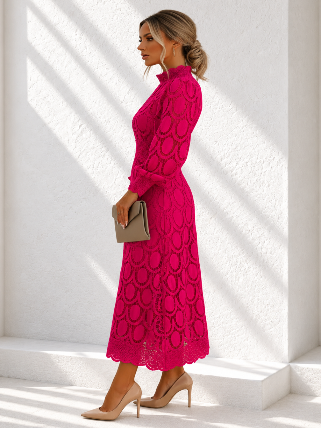 A woman wearing a Villa Vibes Jasmine bright pink lace midi dress with a mock neck and long sleeves, holding a taupe clutch and wearing beige heels, standing in a bright white architectural setting with diagonal shadows.