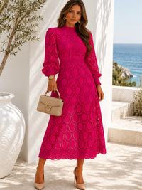 A woman models a Villa Vibes Jasmine bright fuchsia pink lace midi dress with long puff sleeves and a high mock neck, standing on a sunlit outdoor terrace with a white wall and sea in the background. She is holding a beige handbag.