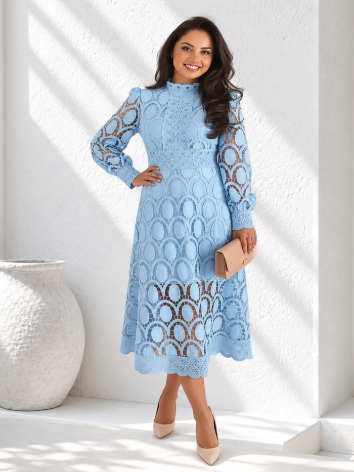 A woman modeling a Villa Vibes Jasmine light blue long-sleeve lace midi dress with a high mock neck and a beige clutch, standing in a bright white room with textured walls and a large white vase.
