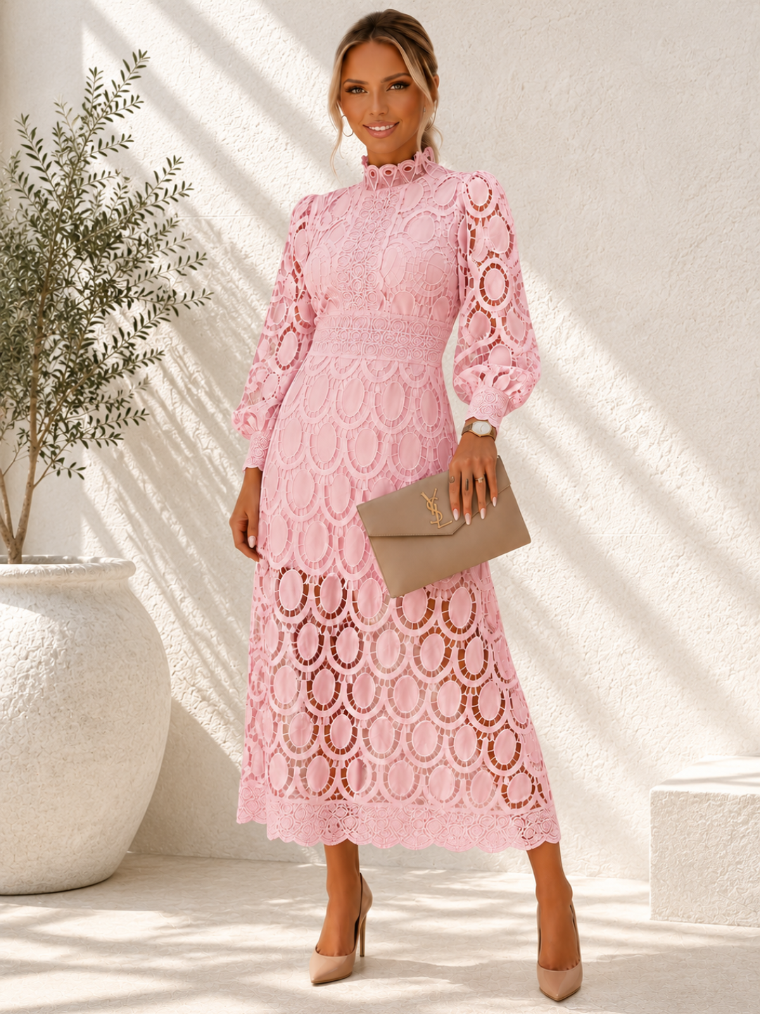 A woman models a Villa Vibes Jasmine  light pink long-sleeve lace midi dress with an allover circular lace pattern, holding a taupe clutch bag and wearing beige heels in a sunlit, textured white room.