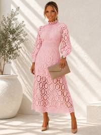 A woman models a Villa Vibes Jasmine  light pink long-sleeve lace midi dress with an allover circular lace pattern, holding a taupe clutch bag and wearing beige heels in a sunlit, textured white room.