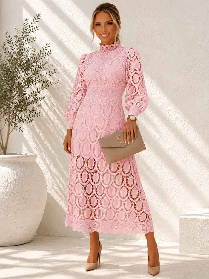 A woman models a Villa Vibes Jasmine  light pink long-sleeve lace midi dress with an allover circular lace pattern, holding a taupe clutch bag and wearing beige heels in a sunlit, textured white room.
