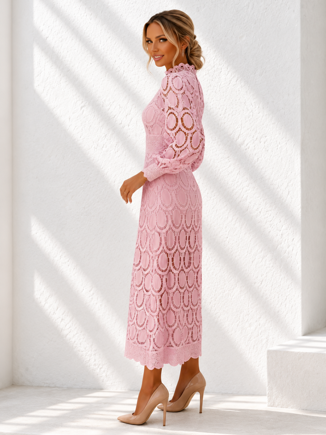 A woman modeling a Villa Vibes Jasmine light pink long-sleeve lace midi dress with a high mock neck and scalloped circular lace pattern, paired with beige high heels, standing in a bright white room with diagonal sunlight shadows.