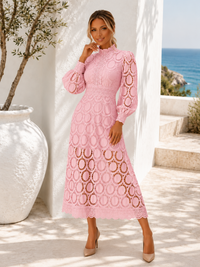 A woman models Villa VIbes Jasmine light pink long-sleeve midi dress with a high mock neck and an allover circular lace pattern, standing in a sunlit outdoor setting with a white wall, large white planter, and ocean view in the background.
