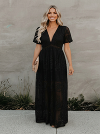 Woman wearing Villa Vibes Lotus black lace dress standing against a gray wall.