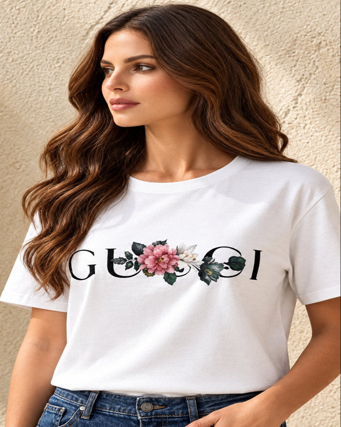 A woman wearing a white short-sleeve t-shirt with a Villa Vibes White GUCCI logo and a pink floral graphic, paired with blue jeans, standing against a textured beige wall.